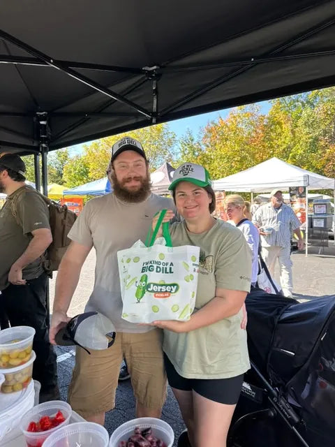 Two customers in Big Dill hats holding a Jersey Pickles tote at the market