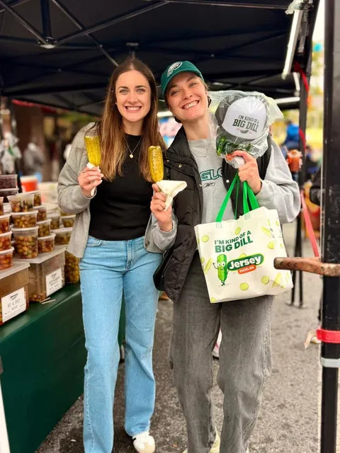 Two customers holding pickles on a stick and a Jersey Pickles tote